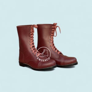 May include: Burgundy leather combat boots with matching laces. The boots have a round toe and a stitched sole. The boots are tall, reaching mid-calf, and are laced up the front. The boots have a logo that says "SWISACO".