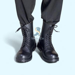 Pode incluir: Um par de botas de combate de couro preto com design de atacadores. As botas t&ecirc;m uma sola resistente e um cano alto, que chega at&eacute; &agrave; panturrilha. As botas s&atilde;o mostradas sobre um fundo azul claro.