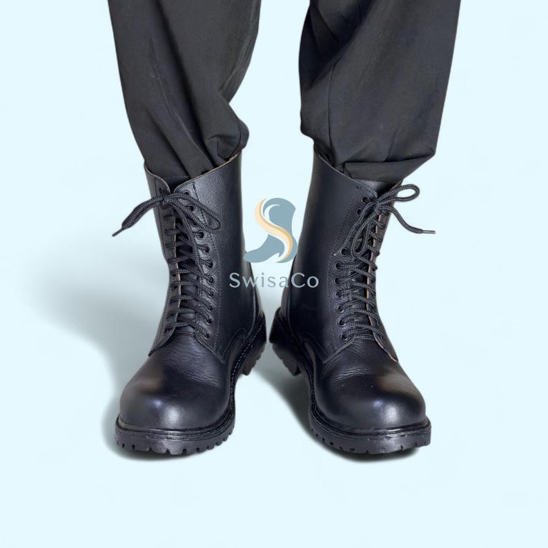 Pode incluir: Um par de botas de combate de couro preto com design de atacadores. As botas t&ecirc;m uma sola resistente e um cano alto, que chega at&eacute; &agrave; panturrilha. As botas s&atilde;o mostradas sobre um fundo azul claro.