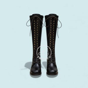K&ouml;nnte beinhalten: Ein Paar schwarze kniehohe Stiefel mit Schn&uuml;rdesign und Messing&ouml;sen. Die Stiefel haben eine runde Spitze und einen braunen Schn&uuml;rverschluss. Die Stiefel werden vor einem hellblauen Hintergrund pr&auml;sentiert.