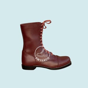 May include: A burgundy leather boot with a cap toe and a lace-up design. The boot has a tall shaft and a black sole with white stitching. The laces are a matching burgundy colour. The boot has the logo "SWISACO" on the side.