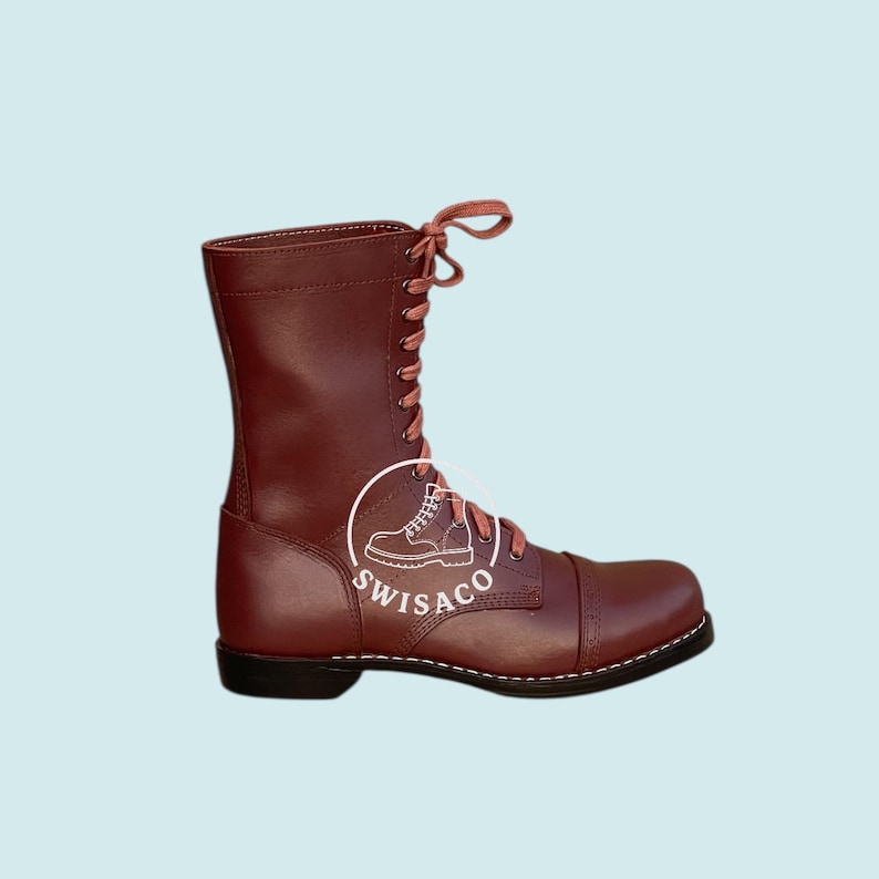 May include: A burgundy leather boot with a cap toe and a lace-up design. The boot has a tall shaft and a black sole with white stitching. The laces are a matching burgundy colour. The boot has the logo "SWISACO" on the side.