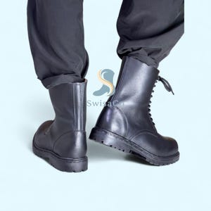 Pode incluir: Botas de combate de couro preto com design de atacadores. As botas t&ecirc;m sola grossa e altura a meio da panturrilha. A imagem mostra as botas de um &acirc;ngulo baixo, destacando sua constru&ccedil;&atilde;o robusta e estilo.
