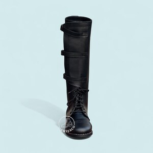 May include: Black knee-high boot with a lace-up front and three buckle straps. The boot has a rounded toe and a tall shaft. The boot is made of a smooth, matte material and is set against a light blue background.