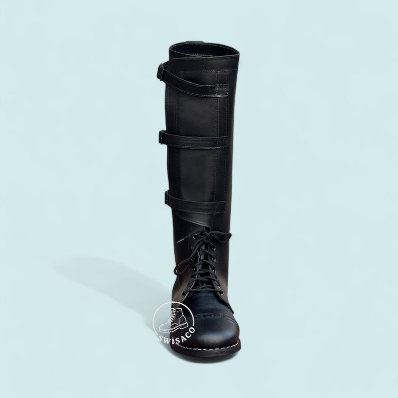 May include: Black knee-high boot with a lace-up front and three buckle straps. The boot has a rounded toe and a tall shaft. The boot is made of a smooth, matte material and is set against a light blue background.