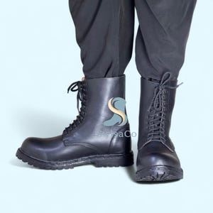 Pode incluir: Botas de combate em couro preto com design de atacadores. As botas t&ecirc;m uma sola resistente e um design de cano alto. As botas s&atilde;o mostradas dos tornozelos para baixo, com o utilizador em cal&ccedil;as pretas. As botas s&atilde;o um estilo cl&aacute;ssico.