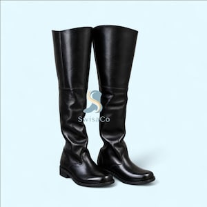 Puede incluir: Un par de botas negras por encima de la rodilla con acabado liso, punteras redondeadas y tacones bajos. Las botas presentan una costura vertical en el lado exterior y un logotipo con el texto SwisaCo situado entre ambas botas.