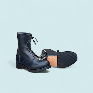 Puede incluir: Botas de combate de cuero negro con diseño de cordones. Las botas tienen una suela resistente y una altura a media pantorrilla. Una bota está de pie, mientras que la otra está tumbada de lado, mostrando la suela. Las botas están sobre un fondo azul claro.