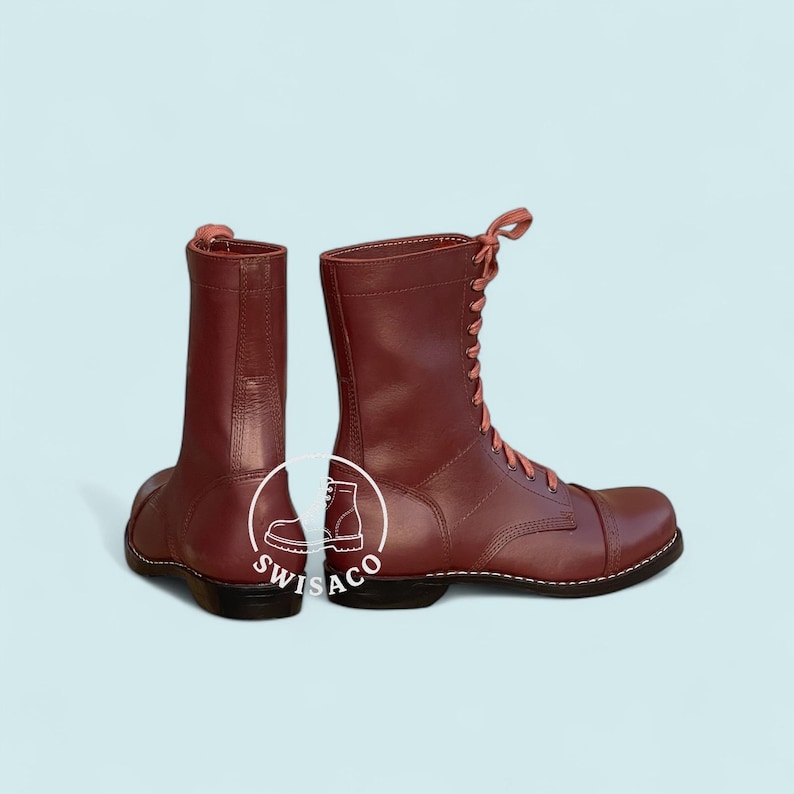 May include: Pair of burgundy leather combat boots with matching laces. The boots have a cap toe design and are set against a light blue background. The boots are a classic style.