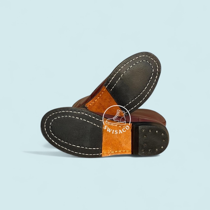 May include: Close-up of the soles of two brown leather boots. The soles are black with white stitching around the edges. The heels are dark brown with small, round studs. A tan leather patch is visible.