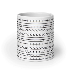May include: White ceramic mug with a repeating black pattern of horizontal lines, zigzags, and other geometric shapes. The mug has a cylindrical shape and a smooth, glossy finish. The design is simple and modern.