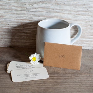 May include: A white ceramic mug with a brown envelope that says "JOY" on it. The envelope is sitting on a wooden surface with a small white daisy flower. The text on the envelope reads: "For His anger is but for a moment, His favor is for life. Weeping may endure for a night, but joy comes in the morning." Psalm 30:5