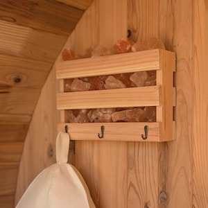 Peut inclure: Support en bois pour pierres de sel de sauna avec trois niveaux remplis de pierres de sel roses et oranges. Le support comporte trois crochets en dessous et est monté sur un mur en bois. Un chapeau de sauna blanc est suspendu à l'un des crochets.