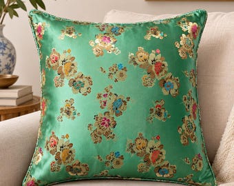May include: A square throw pillow featuring a vibrant green satin fabric with a repeating floral pattern in shades of gold, pink, blue, and cream. The pillow has a matching green piped edge and a smooth, reflective finish across the entire surface.