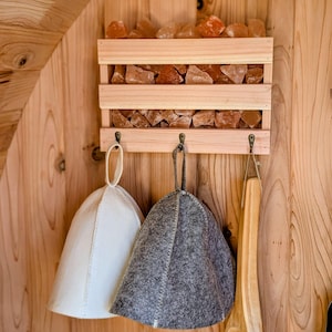 May include: A wooden sauna interior featuring a shelf filled with orange-brown salt rocks. Below, three hooks hold a white and a grey felt sauna hat, and a wooden sauna brush. The wooden structure has a natural grain.