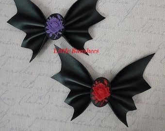 Könnte beinhalten: Zwei schwarze Leder-Fledermausflügel-Haarbänder mit Rosenverzierung. Das obere Band hat eine lila Rose und das untere Band eine rote Rose. Der Text "Little Baushhees" ist im Bild sichtbar.