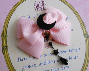 May include: A pink satin bow with a black crescent moon charm and a black beaded chain with star charms and a black teardrop charm. The bow is on a white background with the text "There is no to learn about being a princess, and there are better than fairy"