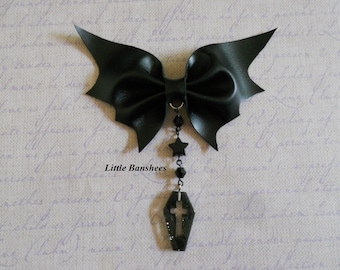 May include: A black bat-wing shaped bow with a dangling charm. The charm features a black star, beads, and a coffin pendant with a cross. The text "Little Banshees" is visible.
