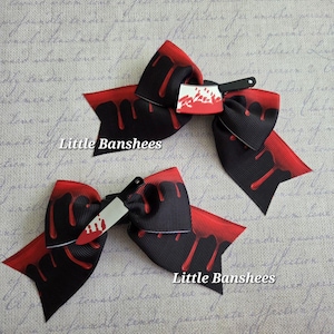 May include: Two black and red hair bows with a dripping blood design and a small knife accessory. The bows have the text "Little Banshees" printed on them. The bows are a Halloween-themed accessory.