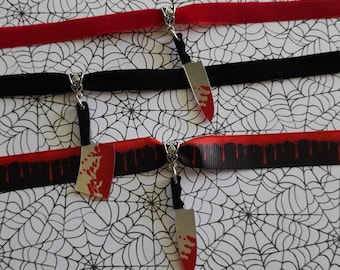 May include: Three chokers with knife pendants. One red velvet choker, one black choker, and one black and red ribbon choker. The knife pendants have a silver finish with red blood splatter details. The background is a white spiderweb pattern.