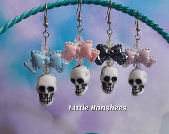 May include: Four pairs of earrings with white skull charms and bow accents in blue, pink, and black. The earrings are suspended from a teal-colored wire. The text "Little Banshees" is displayed above and below the earrings.