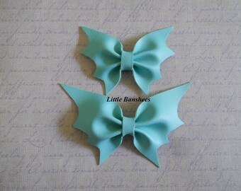 Könnte beinhalten: Zwei hellblaue Leder-Haarschleifen. Jede Schleife hat ein fledermausflügelartiges Design. Die Schleifen befinden sich auf einem strukturierten Hintergrund. Der Text "Little Banshees" ist auf dem Bild sichtbar.