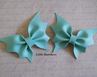 May include: Two light blue leather hair bows in a bat wing shape. The bows are made of a textured material and have a small band in the center. The bows are on a light purple background with the text "Little Banshees".