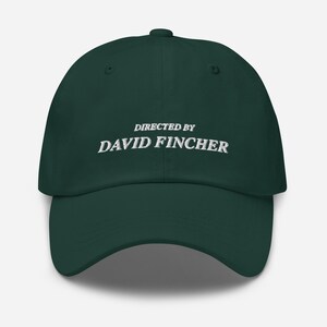 Könnte beinhalten: Dunkelgrüne Baseballkappe mit dem weißen, gestickten Text „DIRECTED BY DAVID FINCHER“. Die Kappe hat einen gebogenen Schirm und einen Knopf oben.