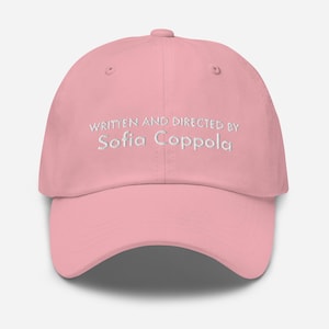 Peut inclure: Casquette de baseball rose clair avec une visière incurvée. Le texte "WRITTEN AND DIRECTED BY Sofia Coppola" est brodé en blanc sur le devant. La casquette a une couronne arrondie et un petit bouton sur le dessus.