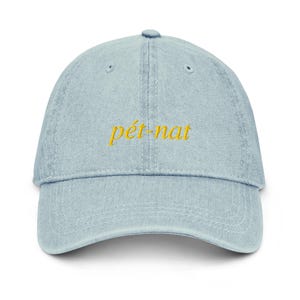 May include: Light blue denim baseball cap with the embroidered text "pét-nat" in yellow. The cap has a curved brim and a fabric strap closure at the back. The hat is a casual accessory.