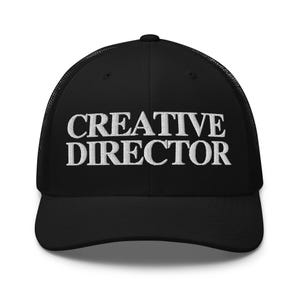 Op de afbeelding: Zwarte truckerpet met de tekst "CREATIVE DIRECTOR" in witte letters geborduurd. De pet heeft een mesh achterkant en een gebogen klep. Een modieus accessoire voor creatieve professionals.