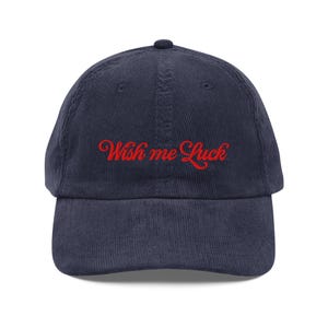Può includere: Cappellino da baseball in velluto a coste blu navy con la scritta ricamata "Wish me Luck" in rosso. Il cappellino ha una visiera curva e un bottone sulla parte superiore. Un accessorio casual.