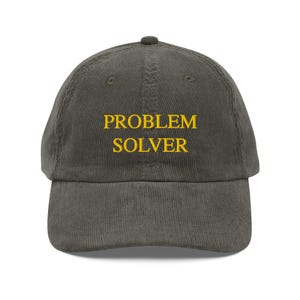Può includere: Cappellino da baseball in velluto a coste verde oliva con la scritta ricamata "PROBLEM SOLVER" in giallo. Il cappellino ha una visiera curva e una parte superiore arrotondata. Il testo è in un carattere grassetto senza grazie.