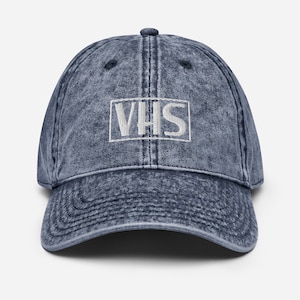 Op de afbeelding: Een denimblauwe baseballcap met een gebogen klep. De cap heeft het woord "VHS" in witte blokletters geborduurd in een witte rechthoek op de voorkant. De hoed heeft een vintage, versleten uitstraling.