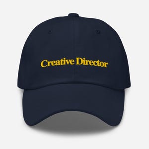 Puede incluir: Gorra de béisbol azul marino con la inscripción bordada "Creative Director" en amarillo. La gorra tiene una visera curva y un diseño clásico de seis paneles. Un accesorio elegante para directores creativos.