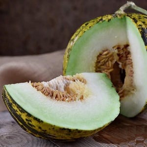 Könnte beinhalten: Eine halbierte Melone mit hellgrünem Inneren und einer Schale mit gelben und schwarzen Streifen. Eine Scheibe der Melone liegt davor und zeigt die Samen und das saftige Fruchtfleisch. Das Obst liegt auf einer Holzoberfläche, was auf ein frisches, natürliches Lebensmittel hindeutet.