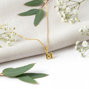 Op de afbeelding: Een goudkleurige ketting met een hanger in de vorm van de letter "M". De hanger is zwart omlijnd. De ketting wordt gepresenteerd op een witte doek met takjes witte bloemen en groene bladeren.