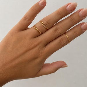 Könnte beinhalten: Ein Set aus drei dünnen Goldringen, die an den Fingern getragen werden. Zwei Ringe sind am Mittelfinger übereinander gestapelt, während ein einzelner dünner Goldring am Ringfinger getragen wird. Die Ringe haben eine glatte, polierte Oberfläche.