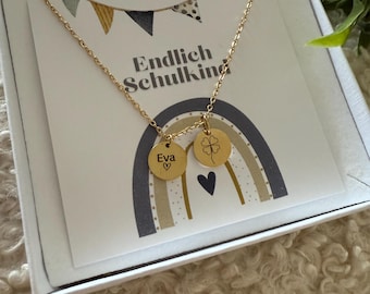 May include: A gold-colored necklace with two round pendants. One pendant has the name "Eva" and a heart, the other has a four-leaf clover. The necklace is presented in a white box with a card that says "Endlich Schulkind" and a rainbow design.
