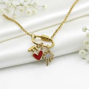 Puede incluir: Collar dorado con cierre tipo mosquetón y cadena. El collar tiene tres colgantes: un corazón rojo, una palmera con cristales transparentes y la letra "F". La joya se exhibe sobre una superficie de satén blanco.