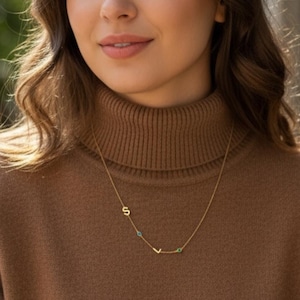 Può includere: Una delicata collana d'oro con le lettere "S" e "L" e piccole pietre preziose turchesi e verdi. La collana è indossata su un dolcevita marrone.