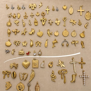Könnte beinhalten: Eine Sammlung goldfarbener Metallanhänger in verschiedenen Formen wie Herzen, Kreuze, Schmetterlinge, Tiere und Symbole. Das Sortiment umfasst kleine Anhänger mit Ösen zur Schmuckherstellung, einige davon mit klaren Steinen oder Perlen verziert.