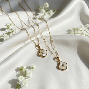 May include: Two gold necklaces with clover-shaped pendants. The pendants feature a white mother-of-pearl inlay with the letters "L" and "P". The necklaces are presented on a white satin surface, with small white flowers arranged around them.