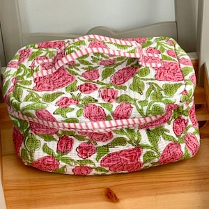 May include: A quilted, rectangular bag with a floral design. The bag has a white base with pink roses and green leaves. The handle and trim are striped pink and white. The bag is on a wooden surface.