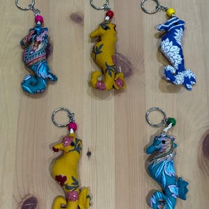 May include: Five fabric seahorse keychains with metal rings. Two are yellow with floral patterns, two are teal with intricate designs, and one is blue with a white mandala-style print. Each features a small coloured bead above the seahorse body.