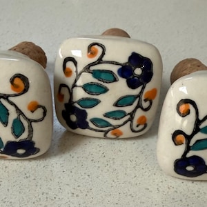 May include: Three square ceramic knobs with rounded corners, each featuring a hand-painted floral pattern in dark blue, teal, and orange on a cream background. Each knob is attached to a small, cylindrical cork stopper base for mounting.