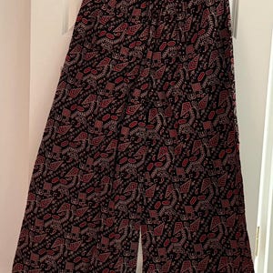 May include: Wide-leg trousers featuring a black base with a repeating pattern of red and white geometric shapes, stylised leaves, and small abstract symbols. The trousers have an elasticated waistband and a loose, flowing silhouette hanging on a hanger.