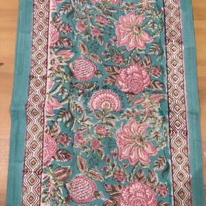 May include: A teal table runner with a floral design in pink and green. The runner has a repeating floral pattern in the centre and a patterned border. The edges are a solid teal colour.