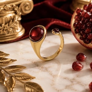 Peut inclure: Une bague ouverte en or ornée de deux pierres cabochons lisses, arrondies et d'un rouge profond de tailles différentes. La plus grande pierre est fixée à une extrémité de l'anneau, tandis que la plus petite est placée à l'autre extrémité.