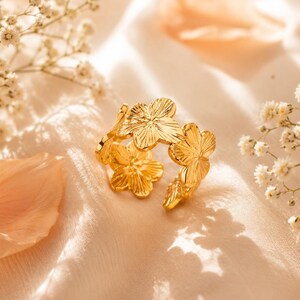 Puede incluir: Un anillo de tono dorado con una banda compuesta por múltiples formas de flores conectadas. Cada flor tiene cinco pétalos con detalles lineales grabados que irradian desde el centro. El anillo tiene un diseño abierto y ajustable.